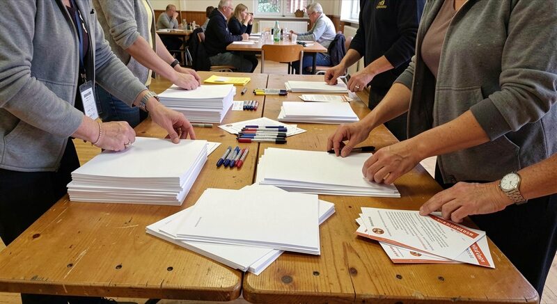 A wooden folding table in a community hall being set with stacked blank sheets, pens and folded paper leaflets, volunteers' hands arranging them